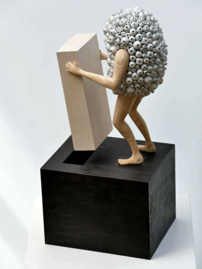Un ajustement profond - François LACOSTE Sculpture en bois : bras et jambes émergeant d'un nuage de perles grises tentant d'insérer une pièce dans une autre
