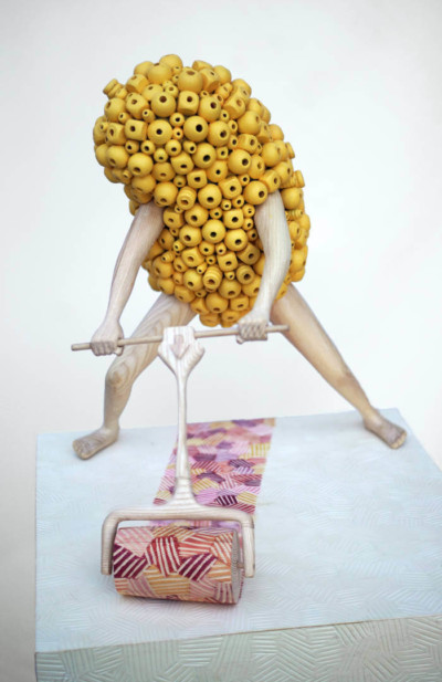 Le pousseur de couleurs - François LACOSTE Sculpture en bois : bras et jambes émergeant d'un nuage de perles jaunes poussant un rouleau encreur